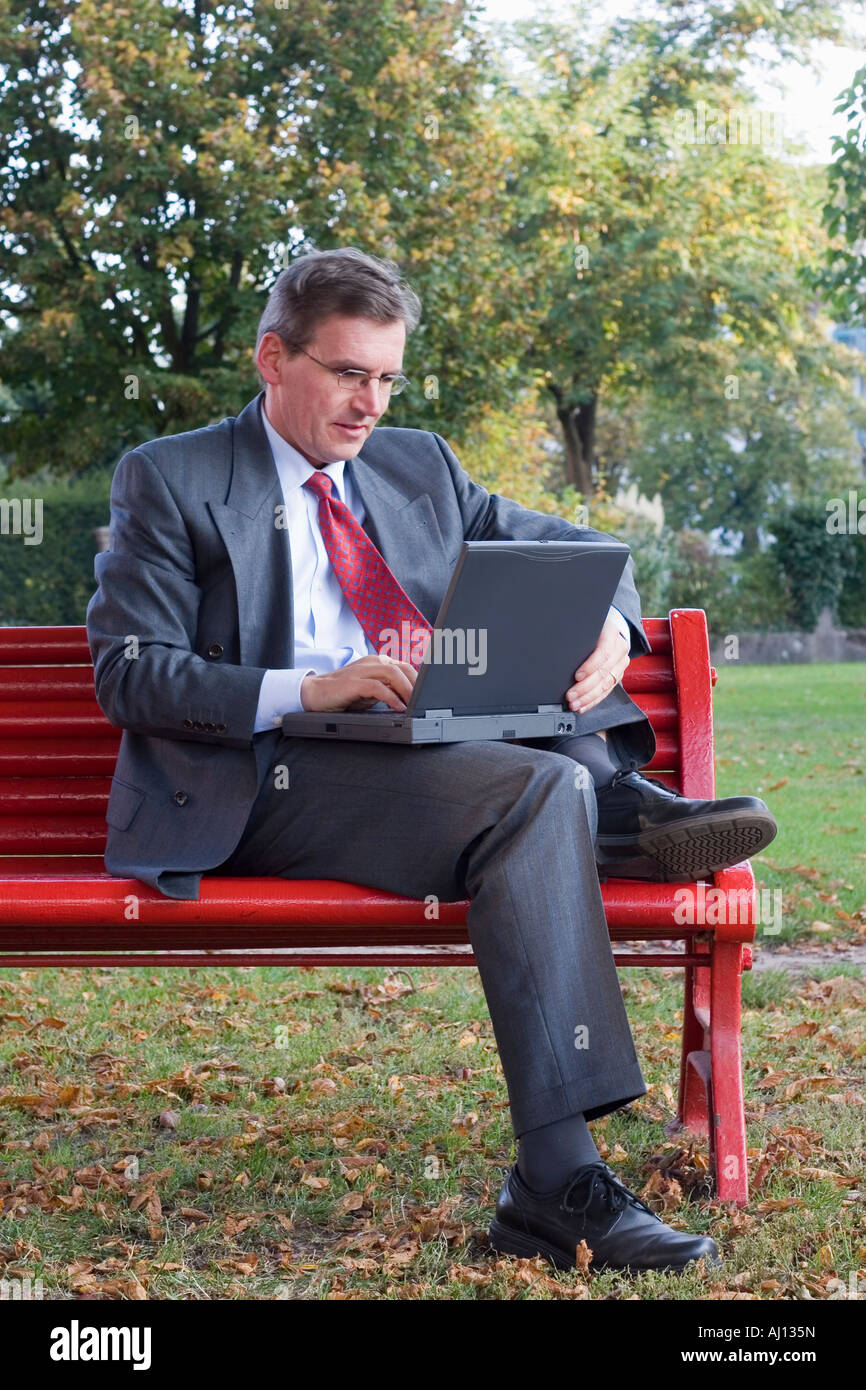 Imprenditore lavora con un computer portatile mentre è seduto su una panchina rossa in un parco Foto Stock