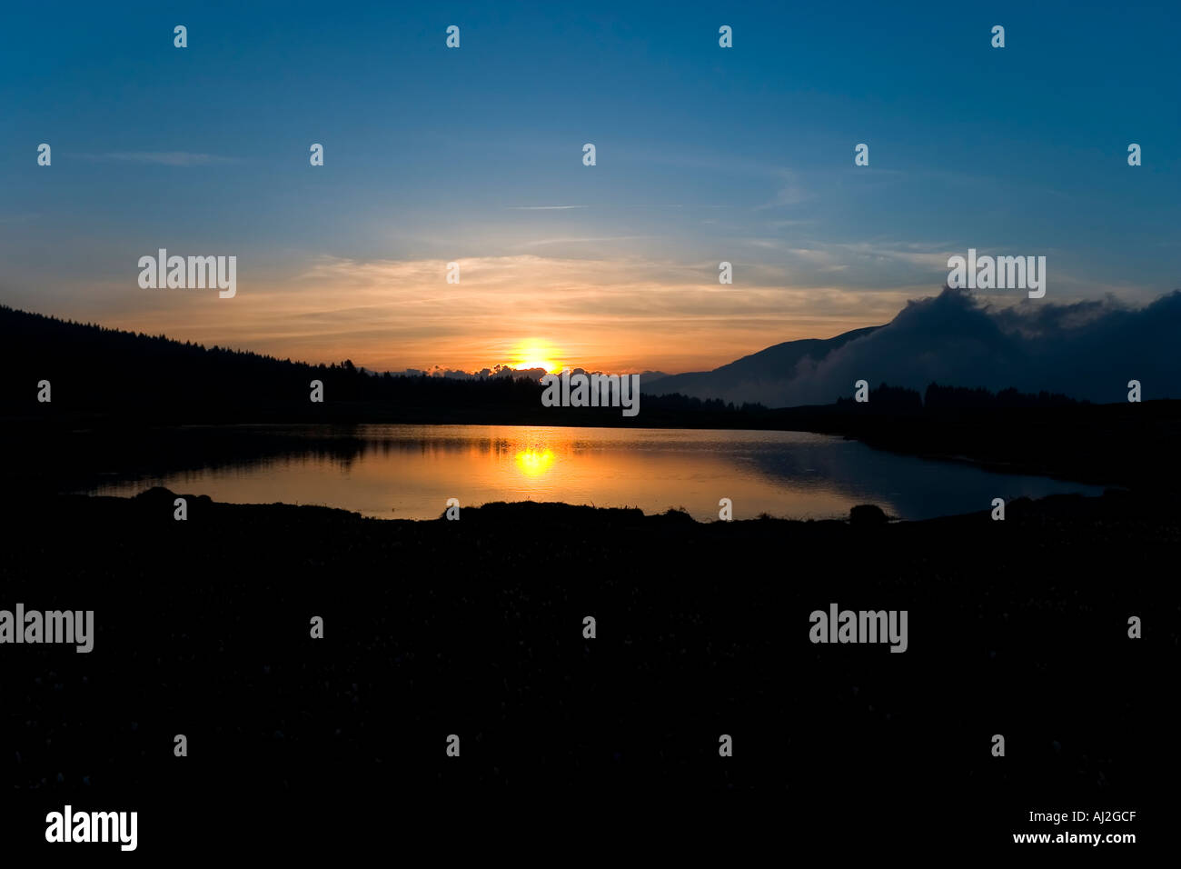 Paesaggio tramonto sul piccolo lago di montagna con il sole rosso che essere specchiata e cielo blu Foto Stock