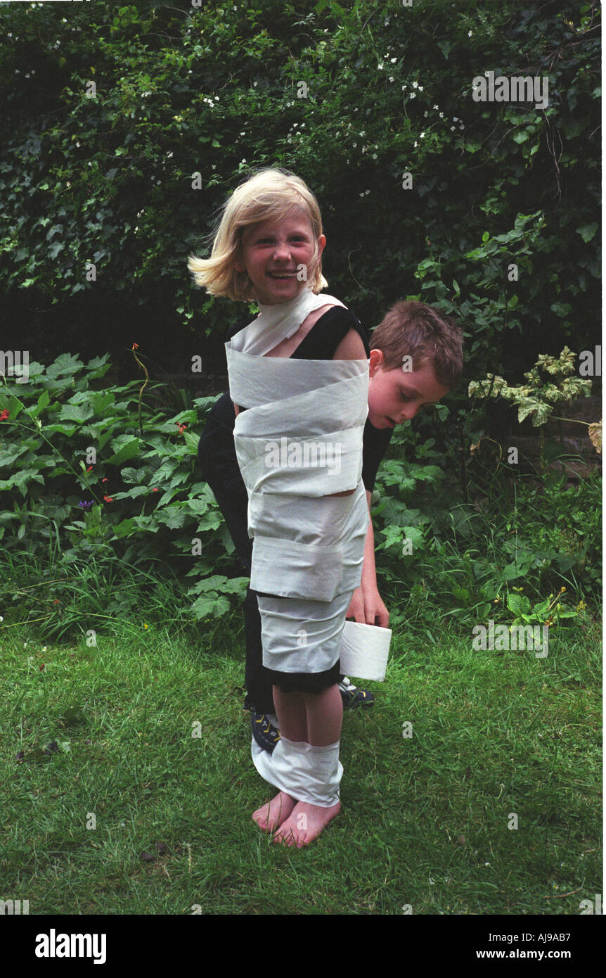 Un ragazzo esegue il wrapping di una ragazza come una mummia egiziana in carta igienica a una festa di compleanno tenutasi in un giardino di Londra, Regno Unito. Foto Stock