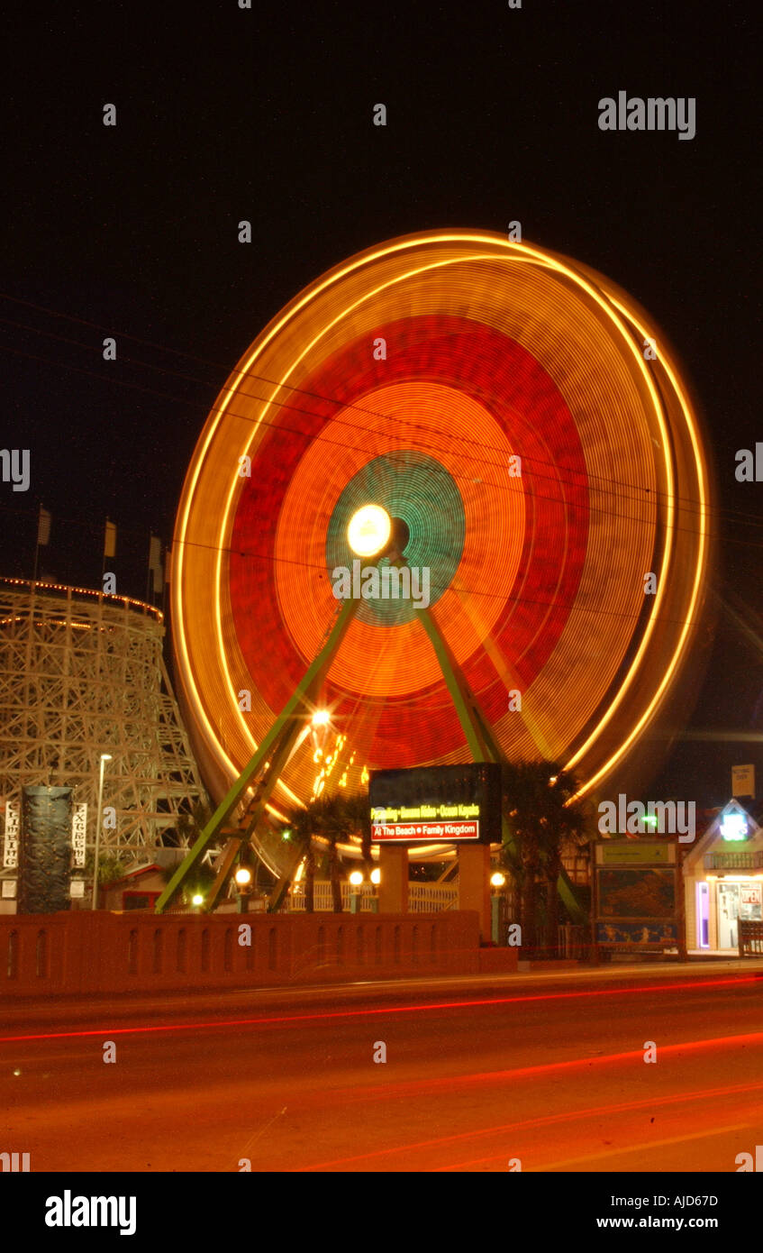 Verticale immagine a colori immagine di una luminosa ruota panoramica Ferris offuscata dalla filatura di notte con le automobili che passa in primo piano Foto Stock