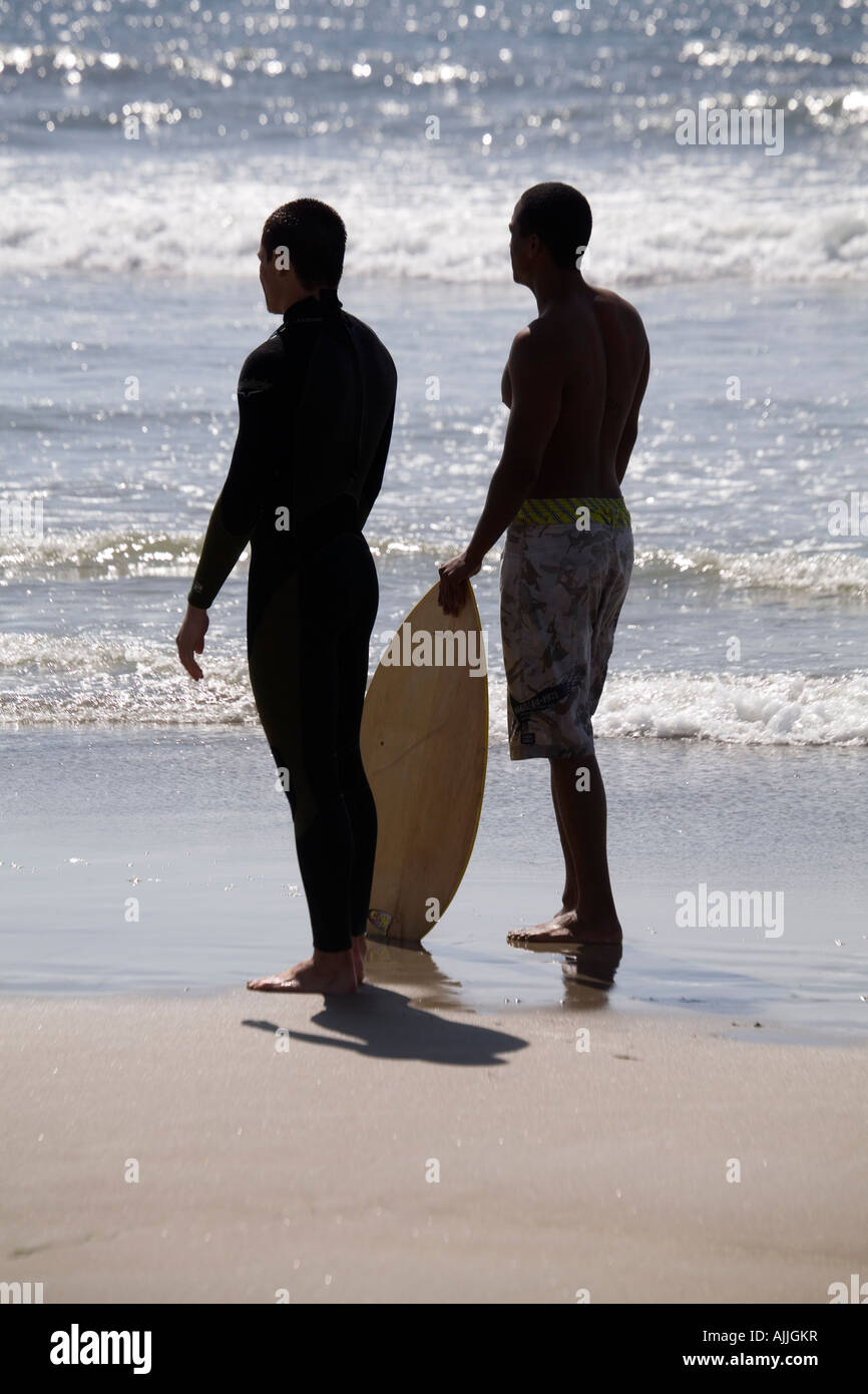 Due giovani uomini riposare su una spiaggia, guardando verso il surf, mentre uno di essi detiene un boogie board. Foto Stock