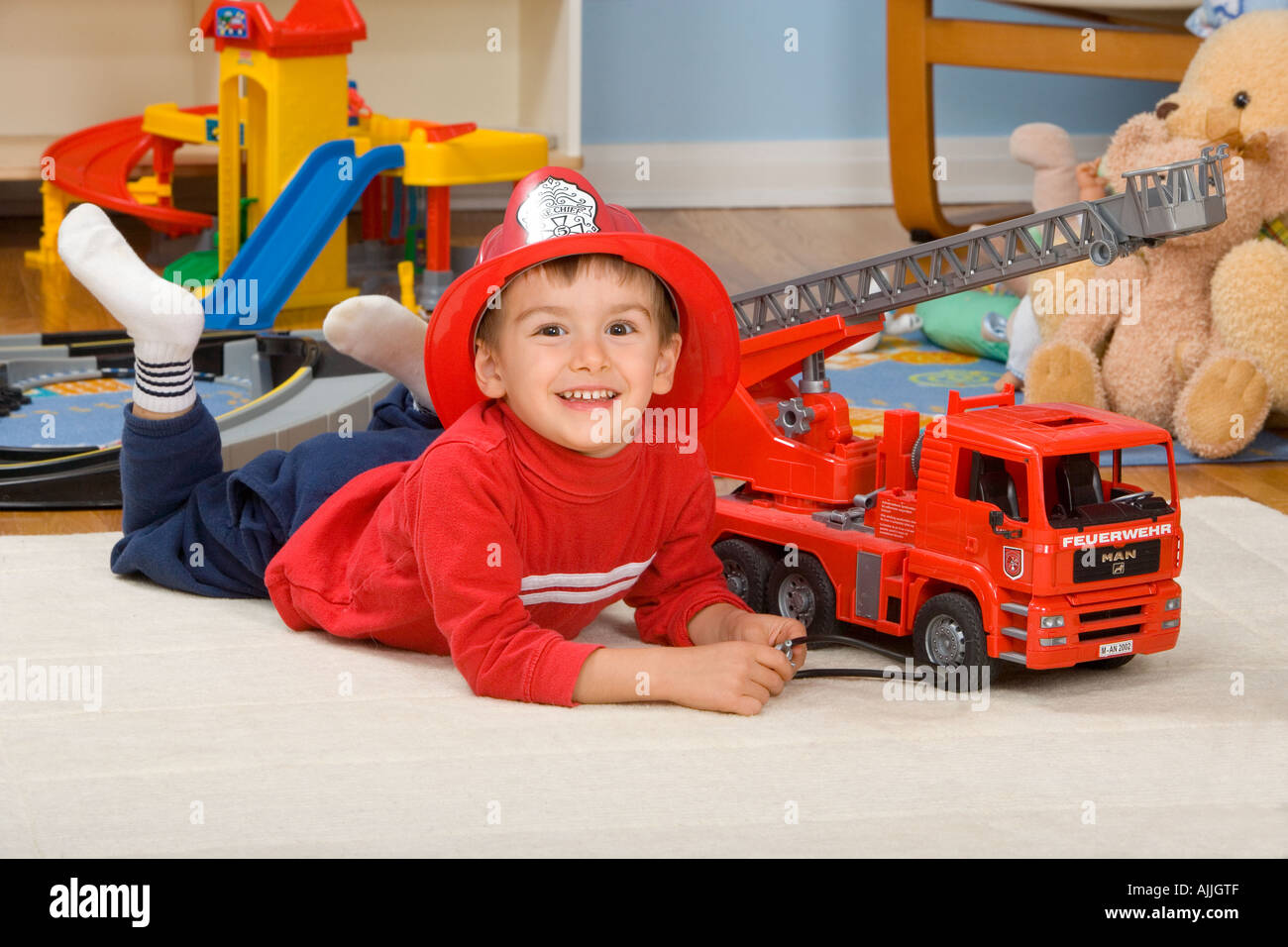 Ritratto di tre anni di vecchio ragazzo che indossa un rosso vigili del fuoco hat seduto accanto a toy red fire carrello nella sua camera da letto Foto Stock