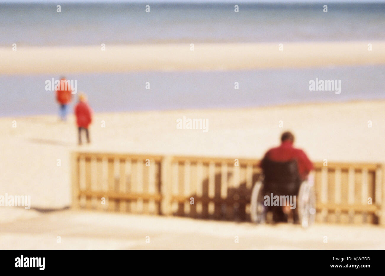 Defocussed persona in sedia a rotelle arrestato in corrispondenza di una barriera sulla spiaggia guardando il mare e a due altre figure Foto Stock