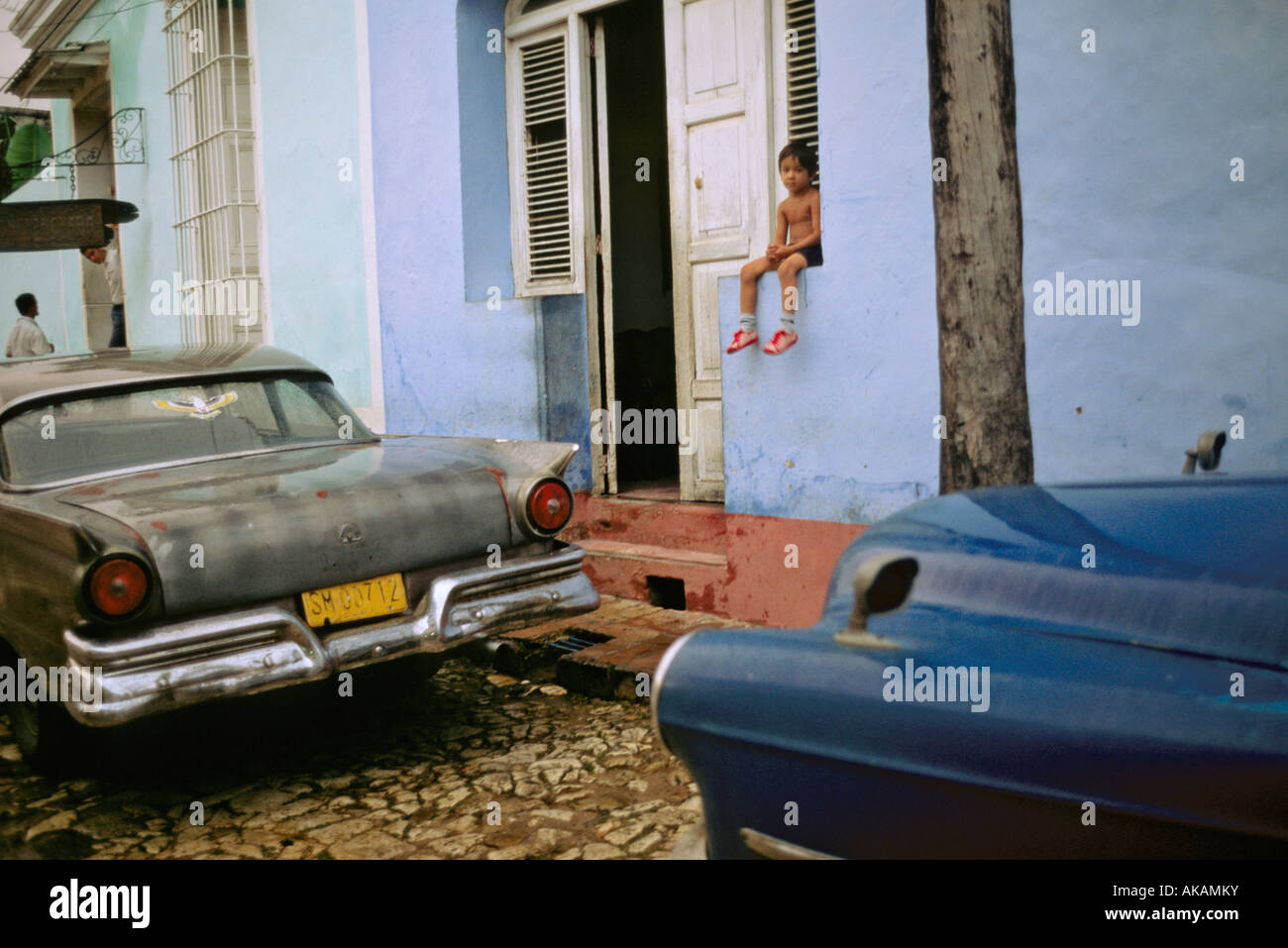 Ragazzo seduto accanto a una strada in ciottoli in Trinidad Cuba tra due vintage auto americane Foto Stock