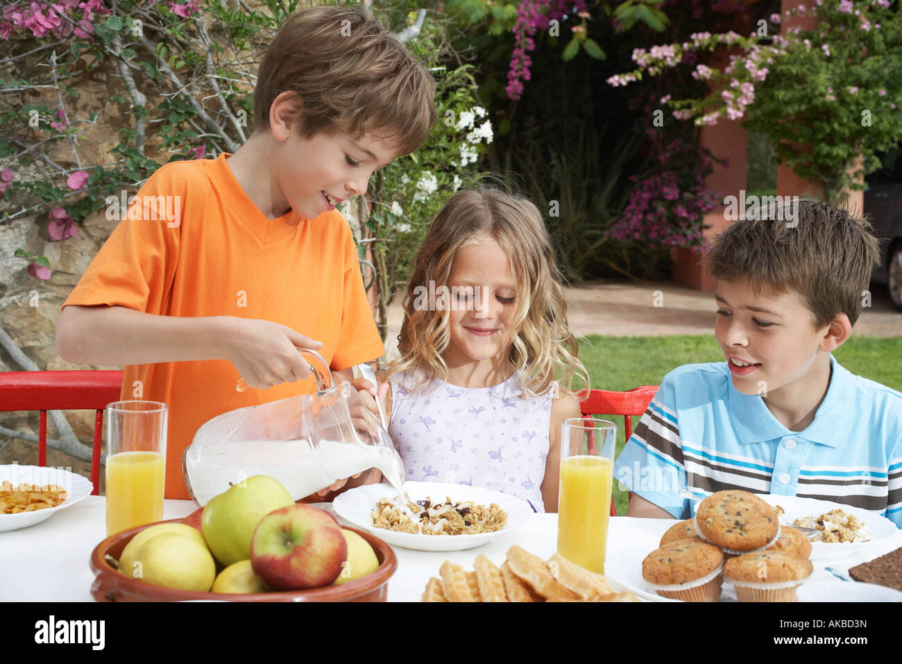 Due ragazzi e una ragazza (6-11) a tavola, ragazzo versando il latte sui cereali, giardino Foto Stock
