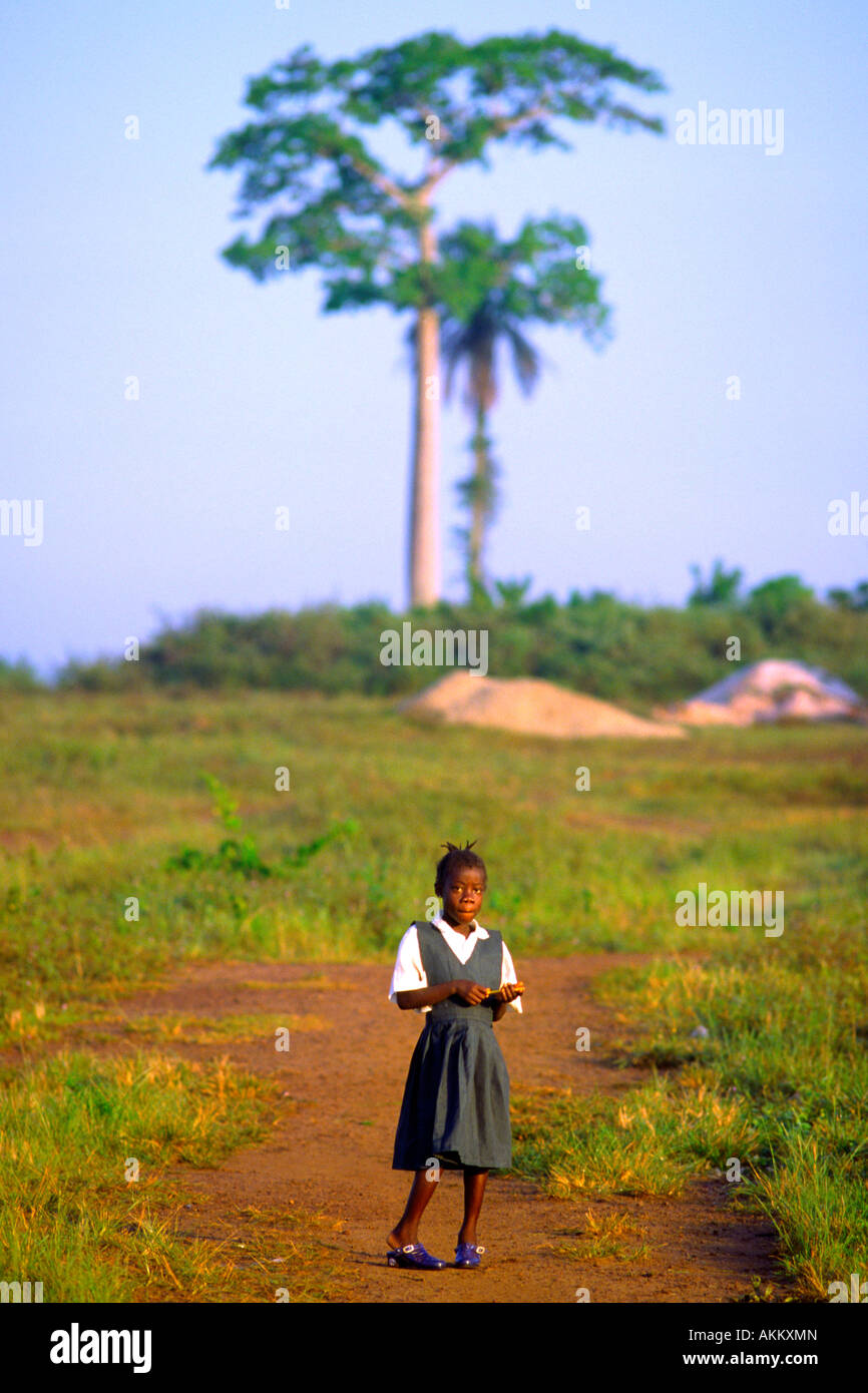 Una ragazza della scuola in uniforme sorge in un campo vuoto con due torreggianti alberi dietro di lei Foto Stock