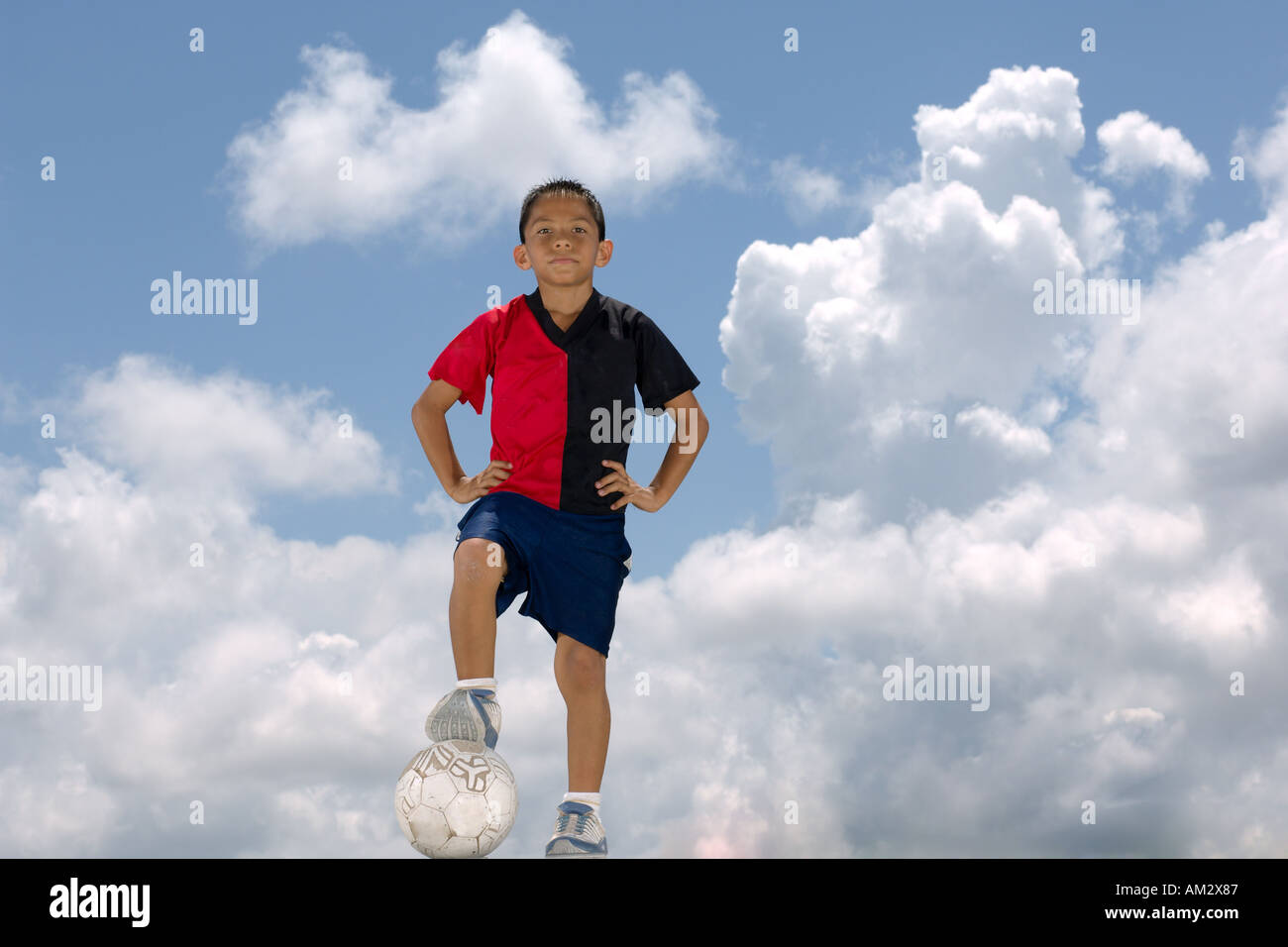 Un ragazzo messicano sta con il calcio di un pallone da calcio di fronte a un cielo blu e nuvole Foto Stock
