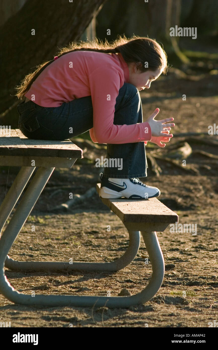 Ragazza russa con una lunga treccia 11 anni seduto sul tavolo nel parco retroilluminata con la luce del tramonto Foto Stock