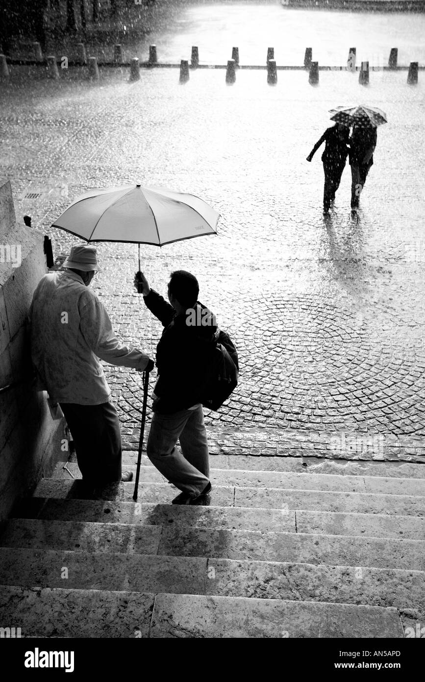 Giovane a piedi sotto la pioggia e il giovane azienda ombrellone a spargere il vecchio uomo Foto Stock