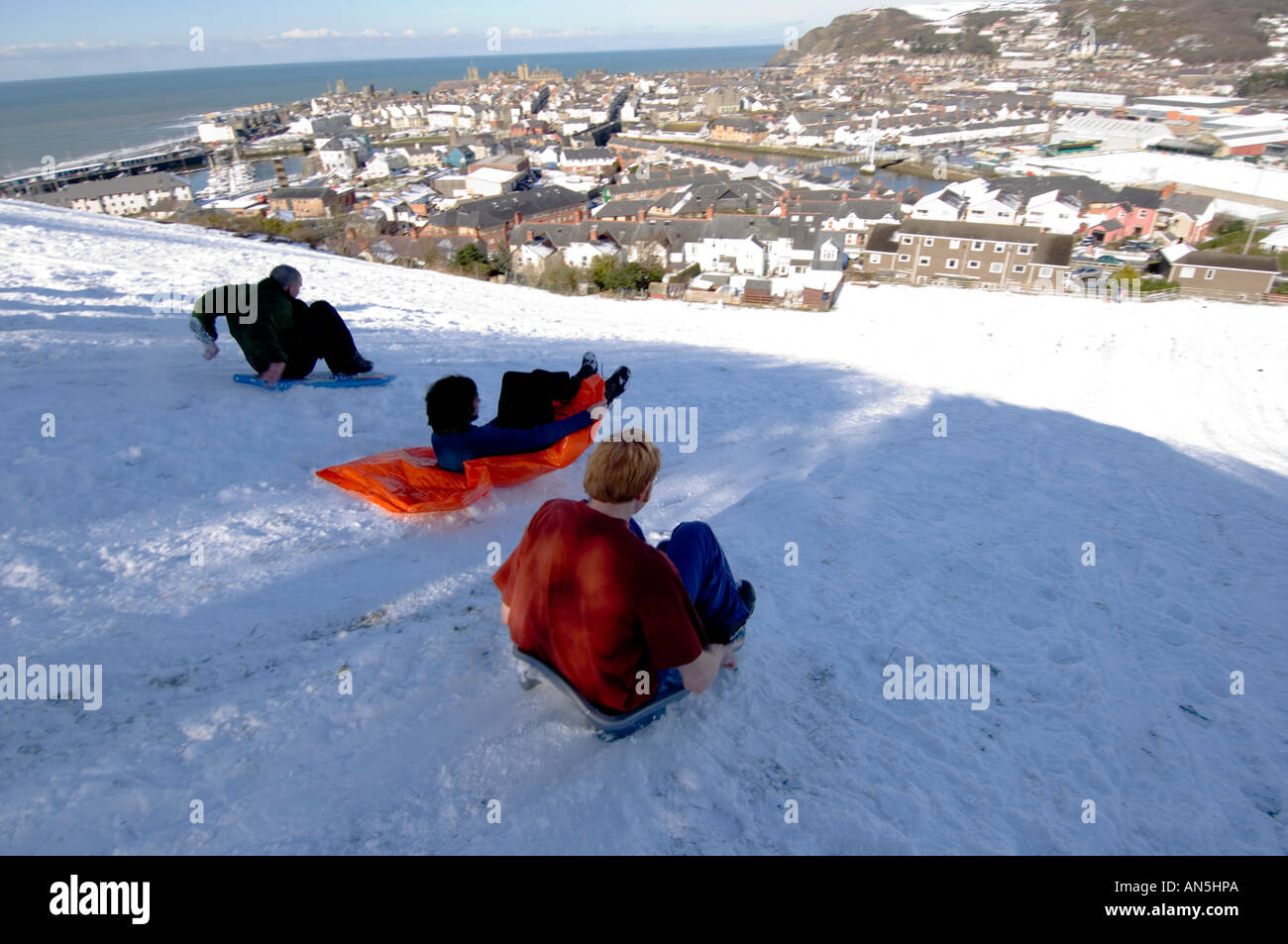 Aberystwyth in inverno la neve - tre ragazzi slittare sulle pendici del pendinas con la città e cardigan bay oltre, Wales UK Foto Stock