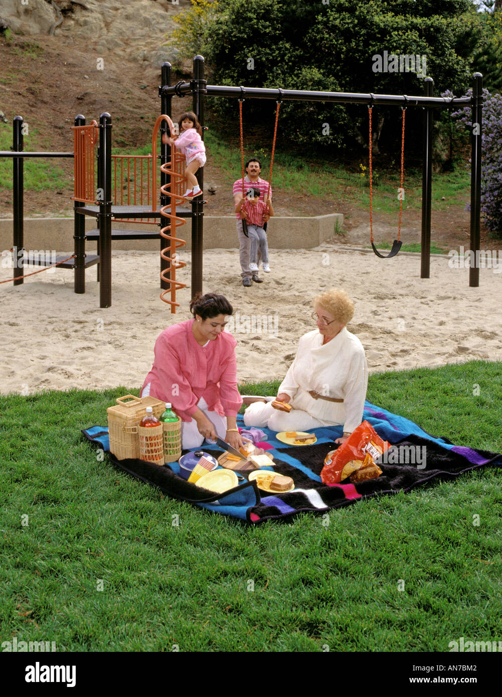 Madre ispanica nonna impostare PICNIC padre mentre i bambini giocano nel parco giochi Foto Stock