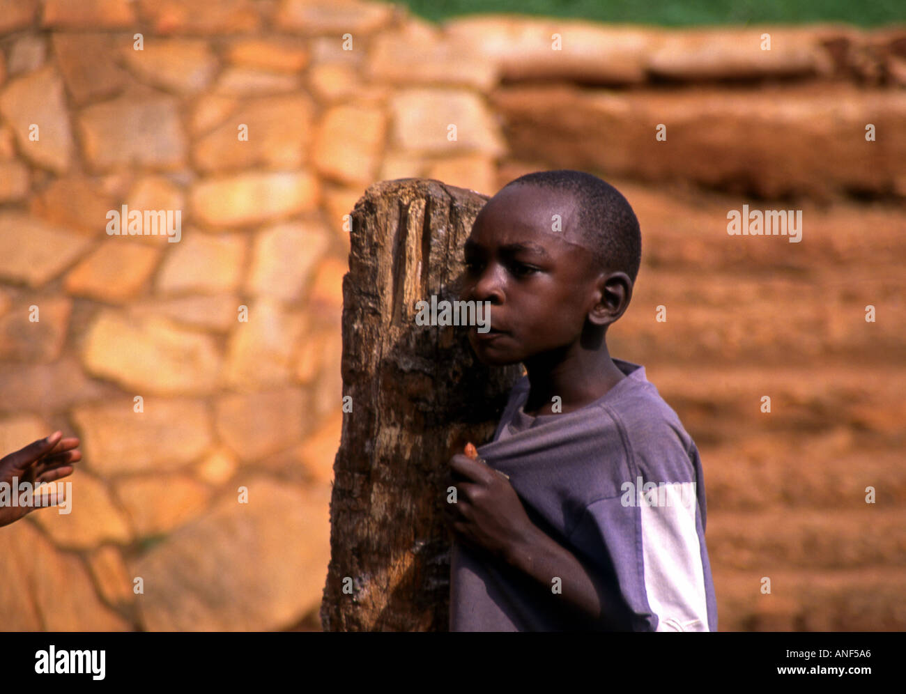 Ritratto bambino ragazzo stand all'aperto in attesa di legno povero post guardare lato soleggiato bright parete in pietra del Jinja Città Uganda Africa orientale Foto Stock
