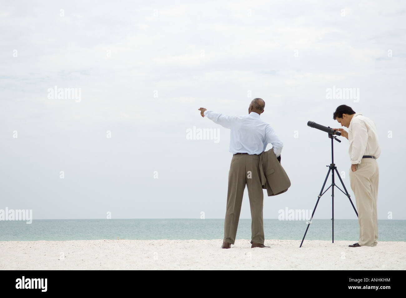 Due imprenditori in piedi presso la spiaggia, uno guardando attraverso il telescopio, l'altra rivolta Foto Stock
