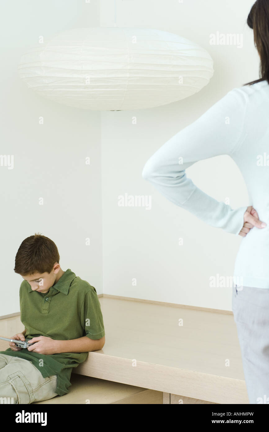 Ragazzo la riproduzione di video gioco, madre a guardare le mani sui fianchi Foto Stock