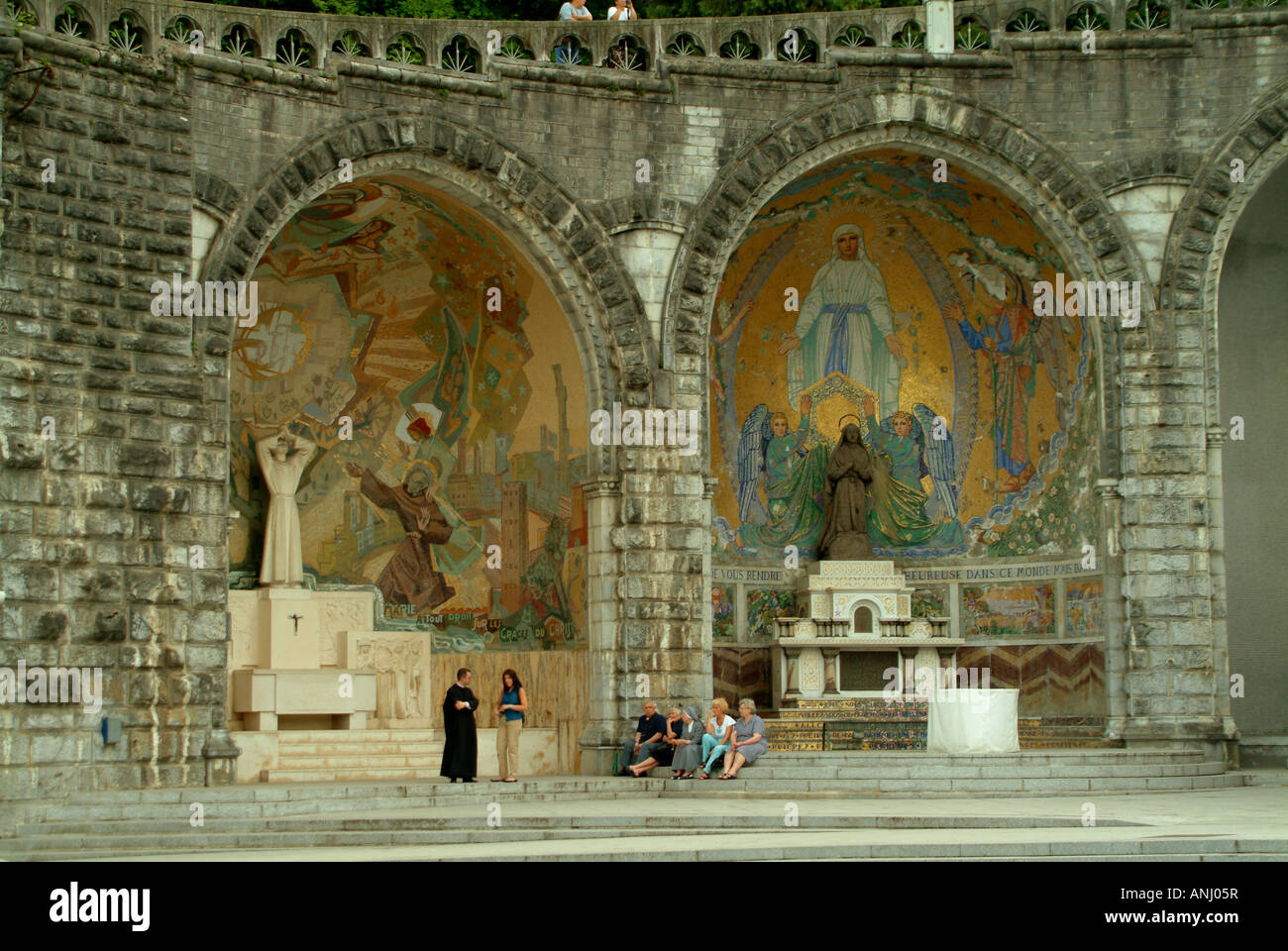 Pellegrini di Lourdes a riposo per una sessione di chat con un sacerdote al di sotto dei mosaici a cupola sotto i passi della basilica Foto Stock