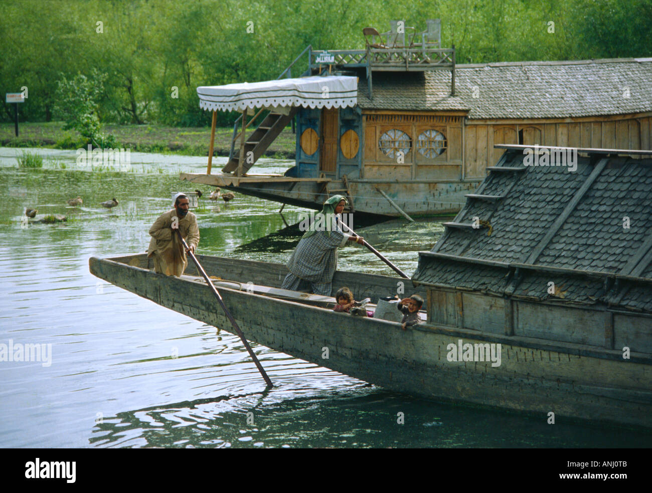 Una famiglia che punisce la loro casa galleggiante tradizionale attraverso il lago dal con una pittoresca casa galleggiante in affitto sullo sfondo. Srinagar, Kashmir. India Foto Stock