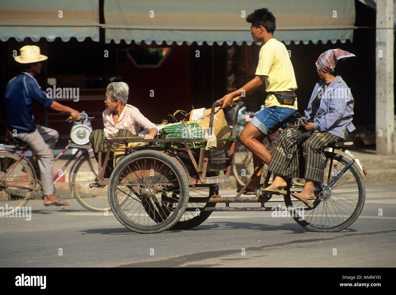 Un uomo in bicicletta a tre ruote di bicicletta carrello con la moglie di equitazione sella laterale sul retro e un'anziana donna seduta sul carrello anteriore accanto a vari fasci su una strada nella città di Sukhothai Foto Stock
