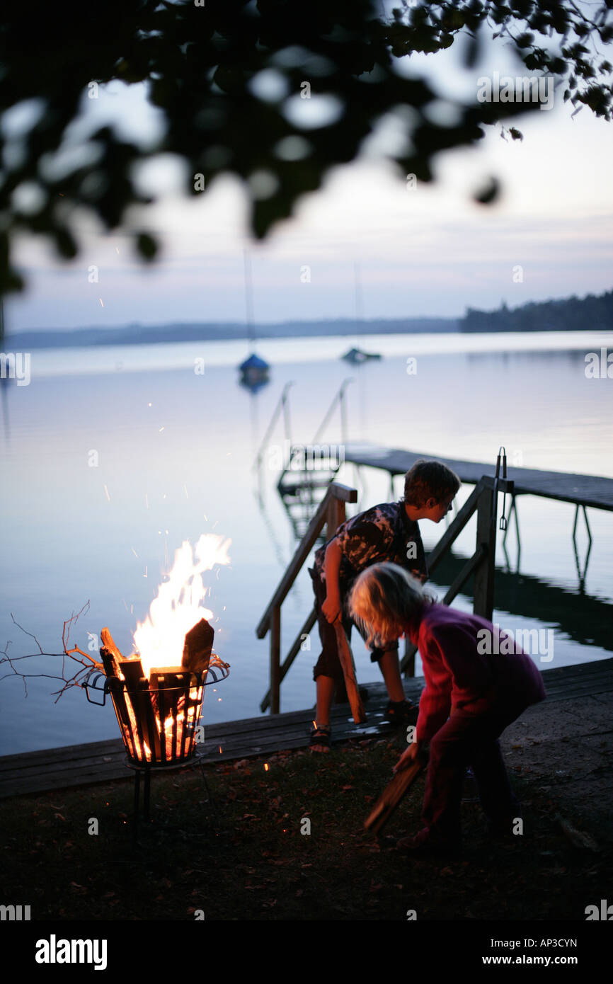 Due bambini in piedi intorno ad un fuoco vicino al molo, sera, Lago Woerthsee, Alta Baviera, Baviera, Germania Foto Stock