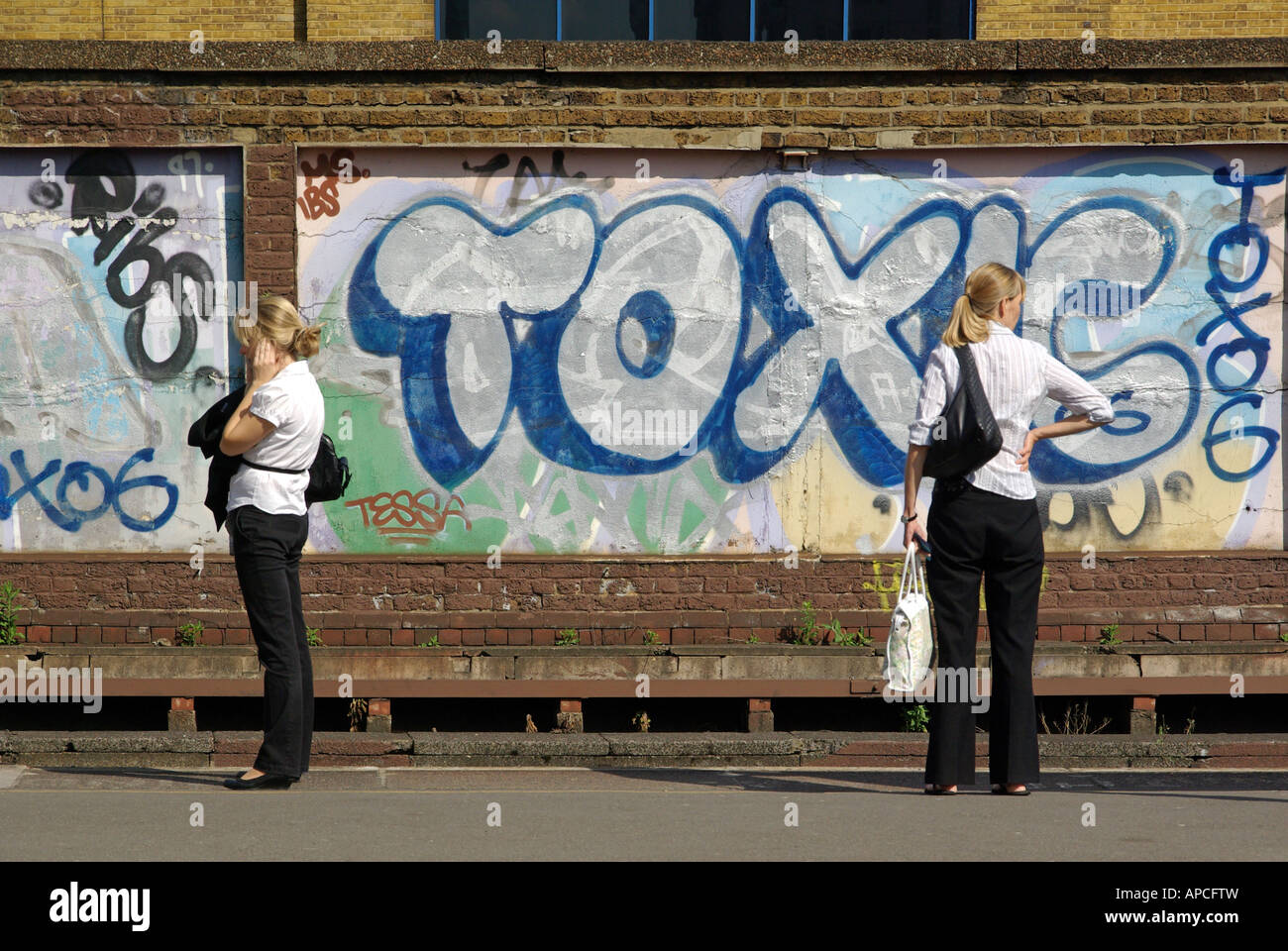 Due donne in piedi all'aperto sulla piattaforma della stazione ferroviaria di South London in attesa di un treno con opere d'arte di graffiti dipinte su pannelli murali Inghilterra Regno Unito Foto Stock