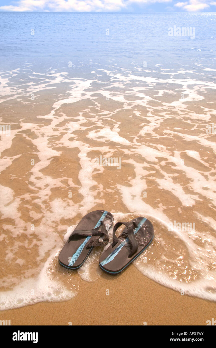 Una coppia di flip flop su una spiaggia di sabbia dorata di essere lavato fuori in mare una leggera sfocatura del movimento Foto Stock