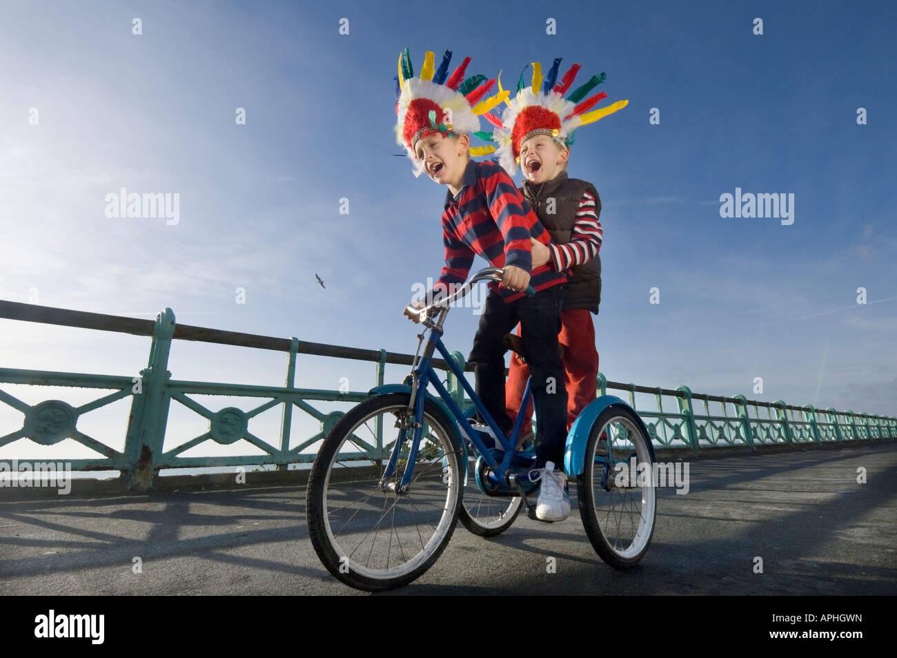 Due bambini piccoli in rosso copricapo indiano urlando come giocano su un trike sulla pista ciclabile sul lungomare di Brighton Foto Stock