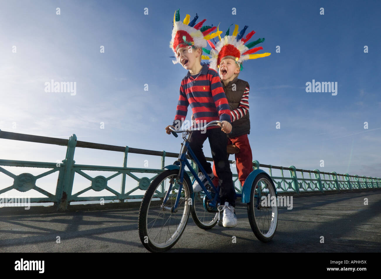 Due bambini piccoli in rosso copricapo indiano urlando come giocano su un trike sulla pista ciclabile sul lungomare di Brighton Foto Stock