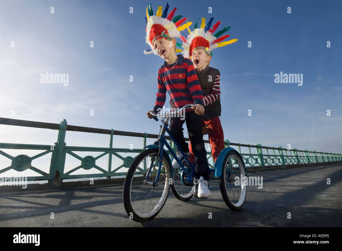 Due bambini piccoli in rosso copricapo indiano urlando come giocano su un trike sulla pista ciclabile sul lungomare di Brighton Foto Stock