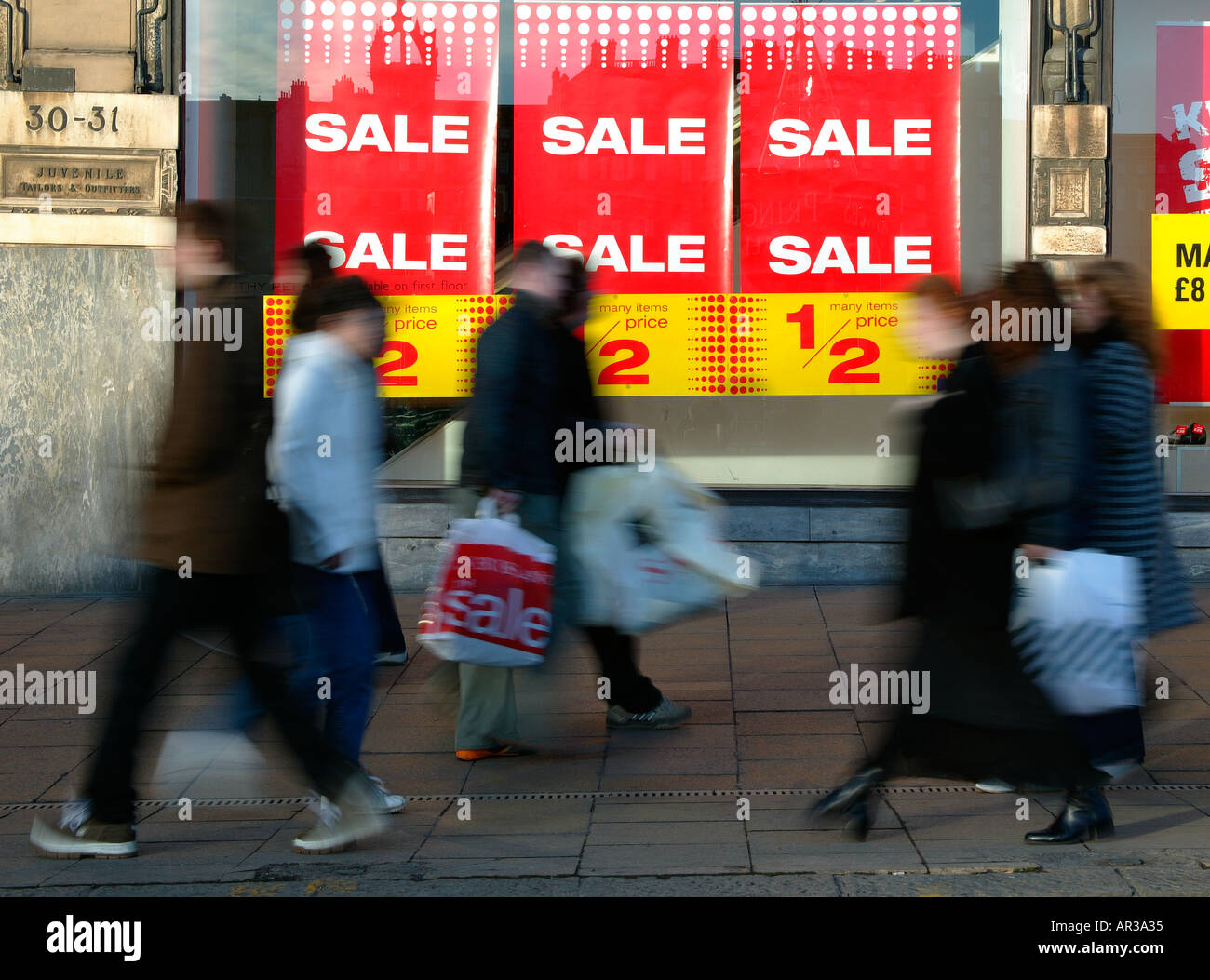 Vendita segni nella finestra di High Street negozio con gli acquirenti in movimento il trasporto del bagaglio in primo piano Foto Stock