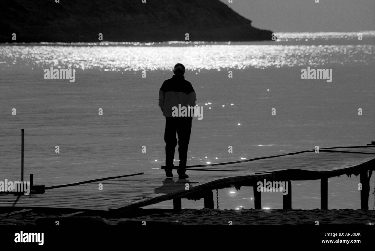 Uomo in silhouette su un molo con vivacemente illuminate dal sole mare sullo sfondo. Foto Stock