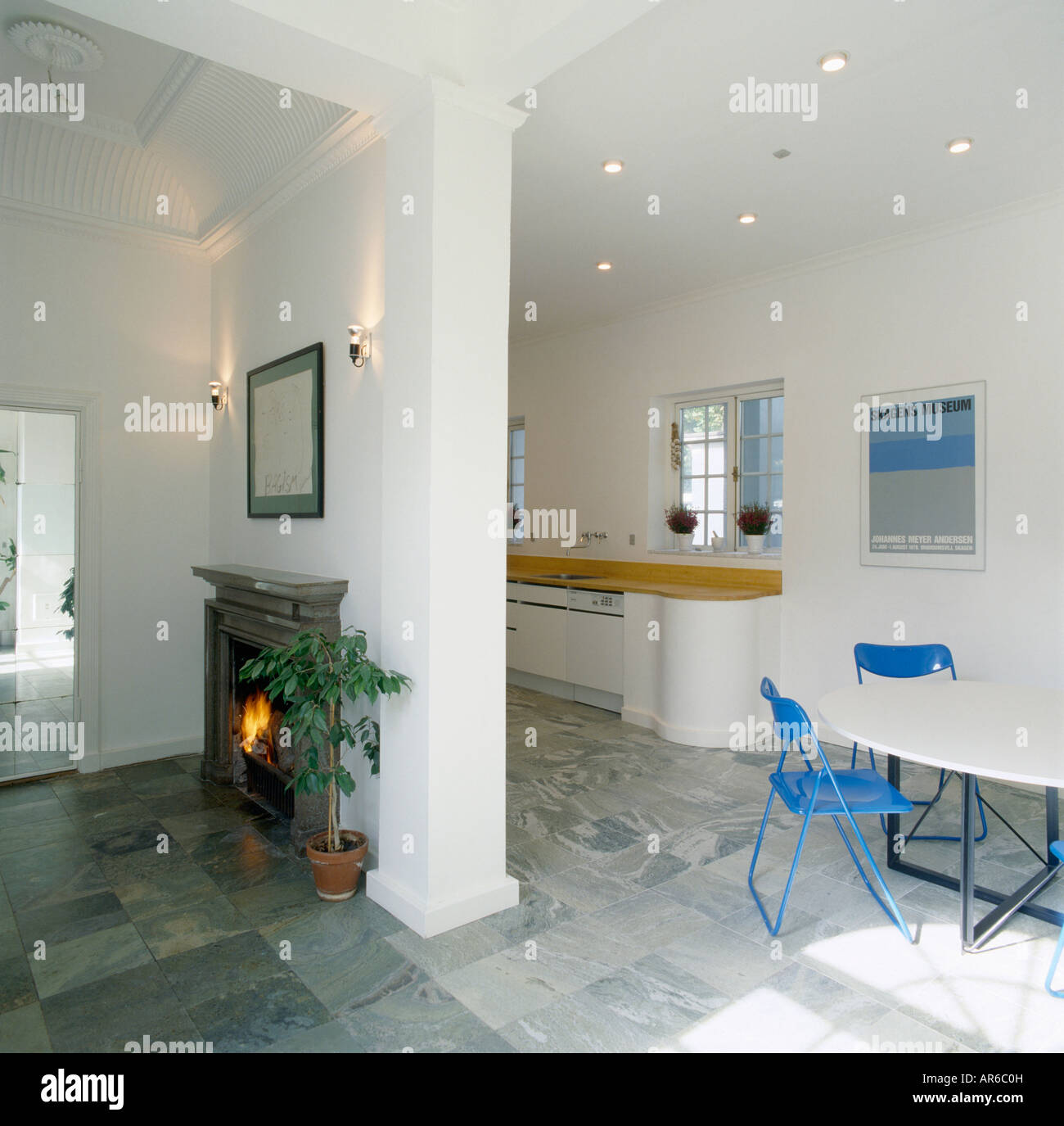 Camino sulla parete divisoria di openplan cucina sala da pranzo Camera con pavimento grigio piastrelle blu e sedie con tavolo bianco Foto Stock