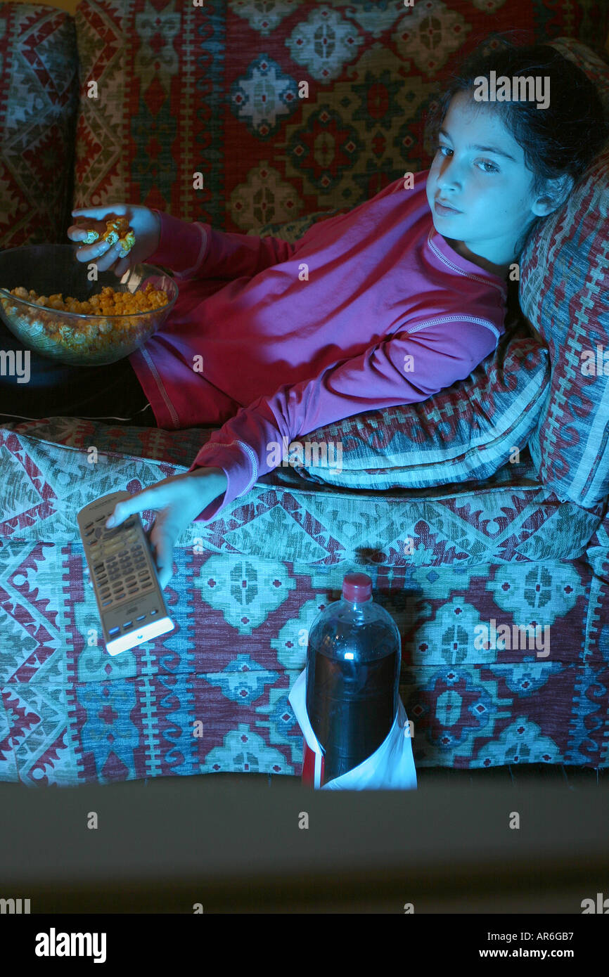 A 10 anno vecchia ragazza (modello rilasciato) mangia pop corn e bevande cola sdraiato sul divano guardando un DVD tenendo premuto il telecomando. Foto Stock