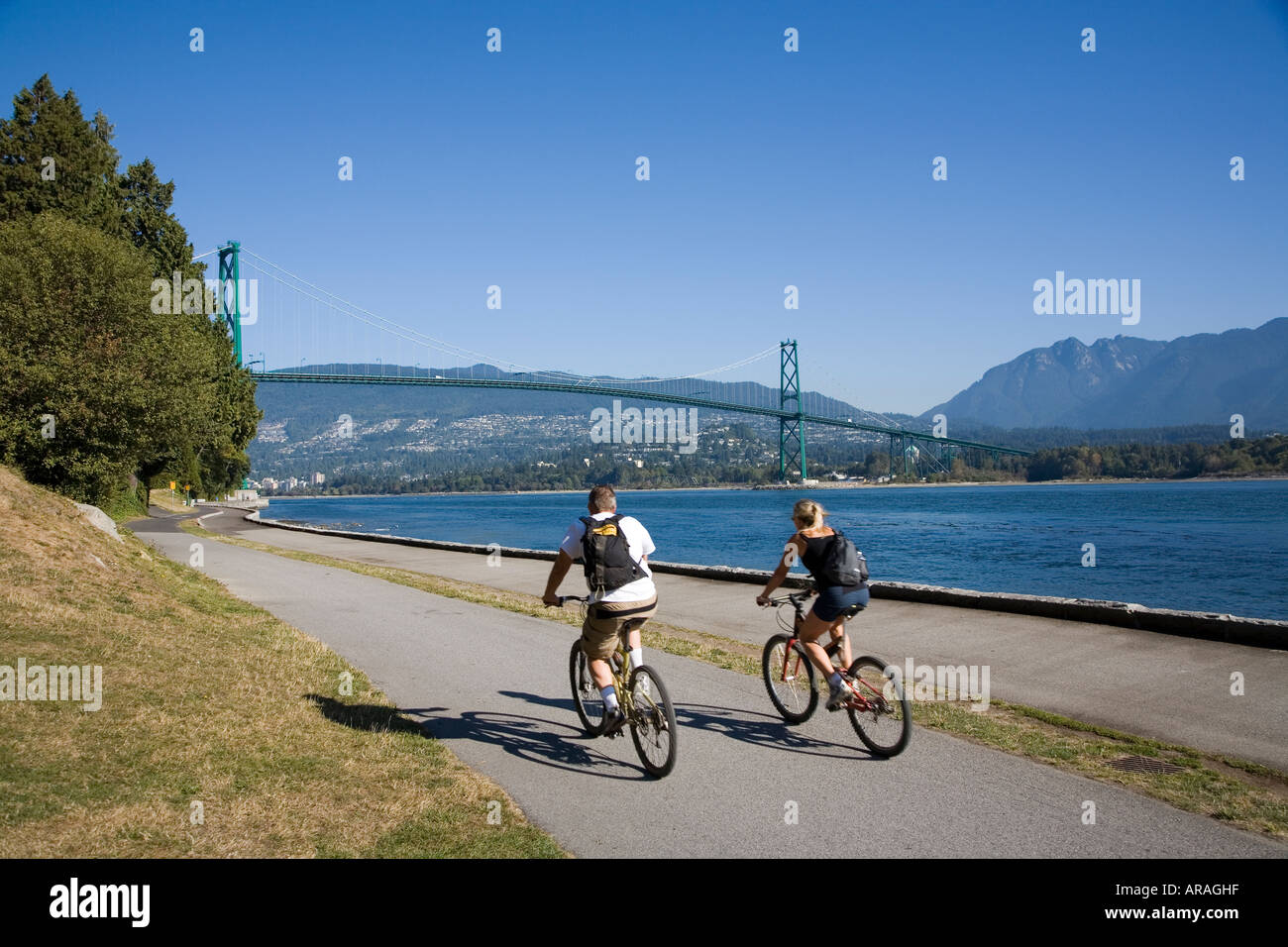 Due ciclisti sulla pista ciclabile nel Parco di Stanley con Ponte Lions Gate in background Vancouver British Columbia Canada Foto Stock