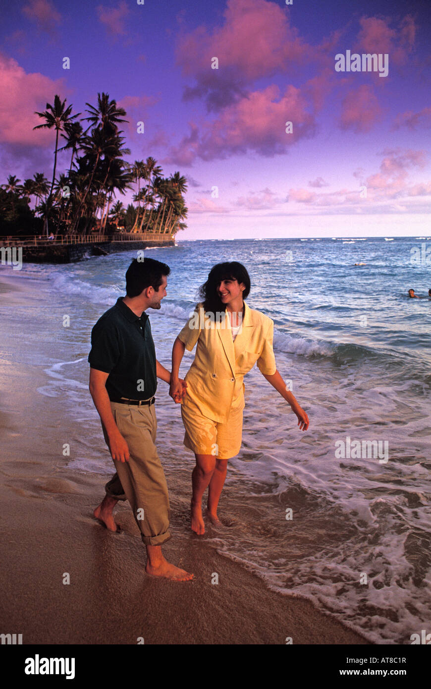 Completamente vestito, barefoot giovane tenendo le mani a piedi lungo l'oceano in prima serata con palme e Nuvole rosa Foto Stock