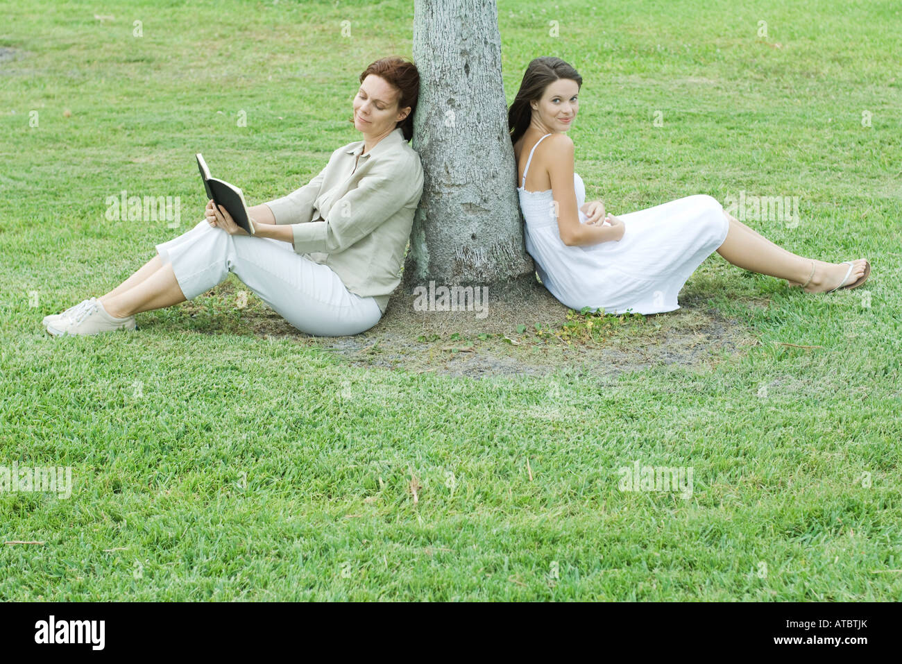 Madre e figlia appoggiata contro il tronco di albero, donna libro, teen ragazza sorridente in telecamera Foto Stock