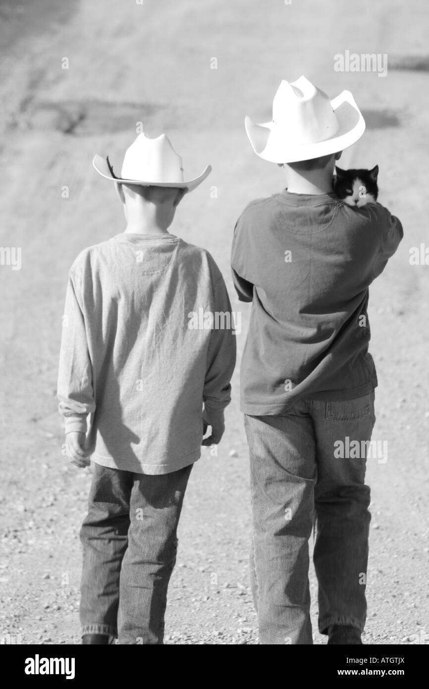 Due ragazzi con i cappelli da cowboy camminare in una fattoria lane con cat on boys' spalla Foto Stock