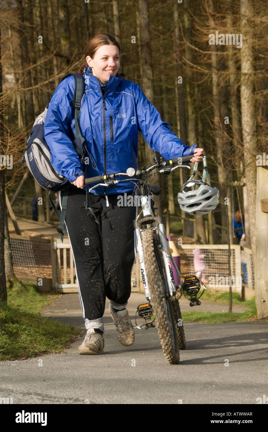 Giovane donna sorridente spingendo la sua bici sul percorso di Nant Yr Arian parco forestale Ceredigion il Galles Centrale dopo un giro nei boschi Foto Stock