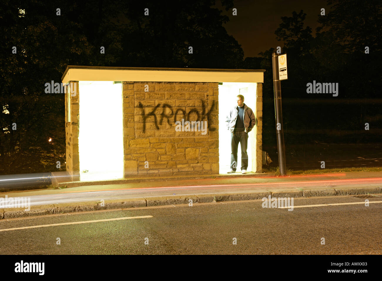 Un uomo alla fermata dell autobus di notte con un blocco a torre in background e una strada in primo piano. Foto Stock