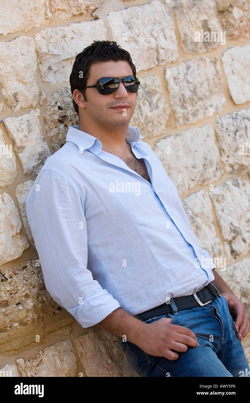 Giovane uomo che indossa gli occhiali da sole in piedi accanto ad una parete le mani sui suoi pantaloni Foto Stock