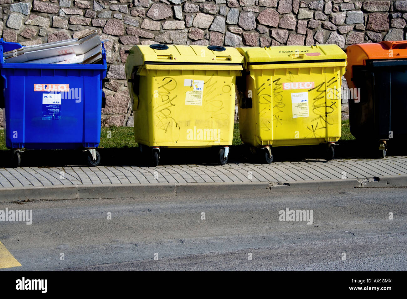 Contenitori di riciclaggio blu, gialli e arancioni per lo smistamento dei rifiuti su un marciapiede pavimentato con un muro di pietra e erba verde sullo sfondo sotto la luce del sole. Foto Stock