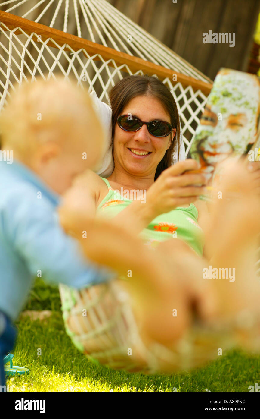 Donna rilassante in un amaca in giardino, leggendo una rivista, piccolo ragazzo accanto a legare l'amaca, relax Foto Stock