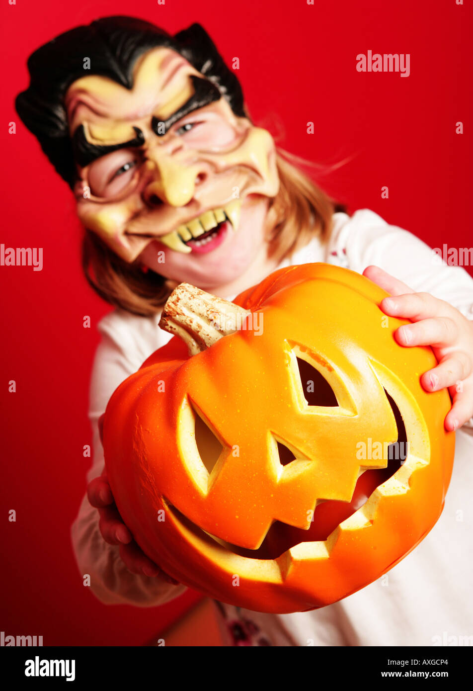 Un singolo giovane ragazza caucasica con maschera vampiro sulla detenzione di un arancione zucca di fronte a lei contro uno sfondo rosso Foto Stock