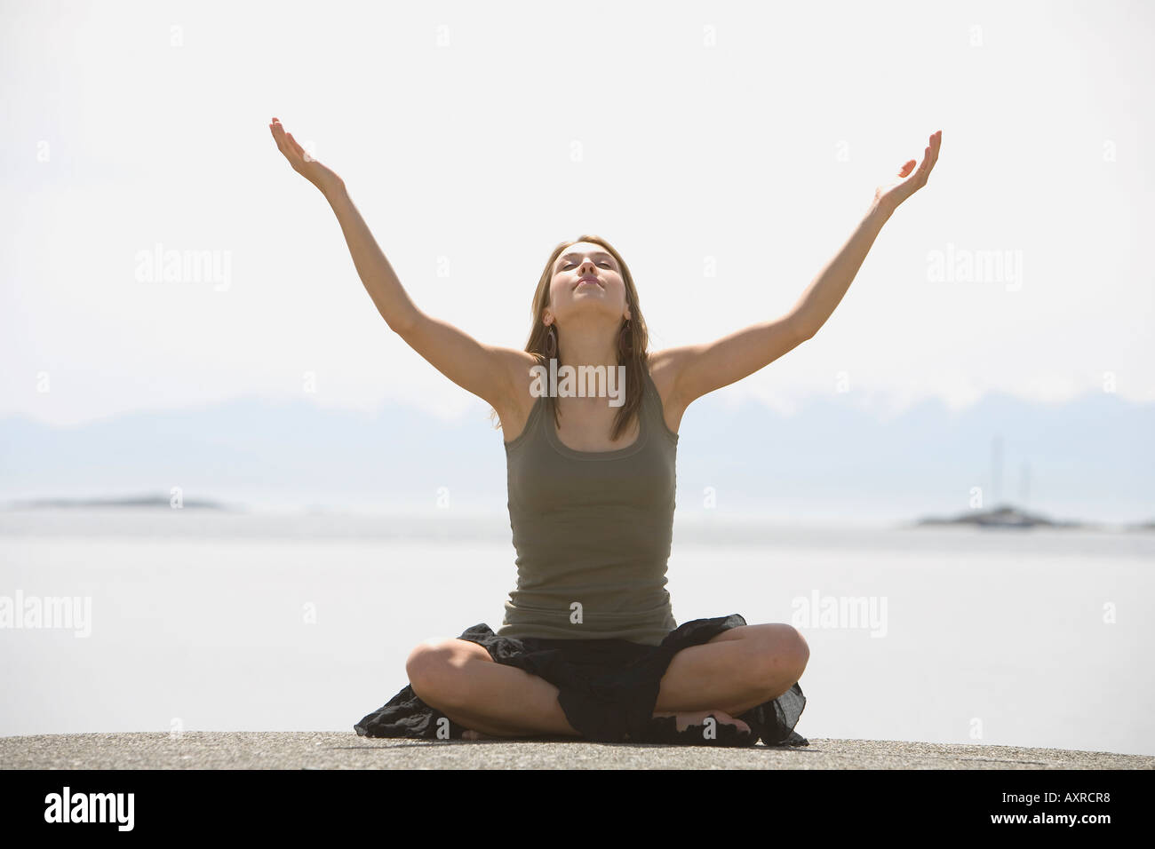 Giovane donna seduta zampe a croce con bracci sollevati di fronte spiaggia Foto Stock