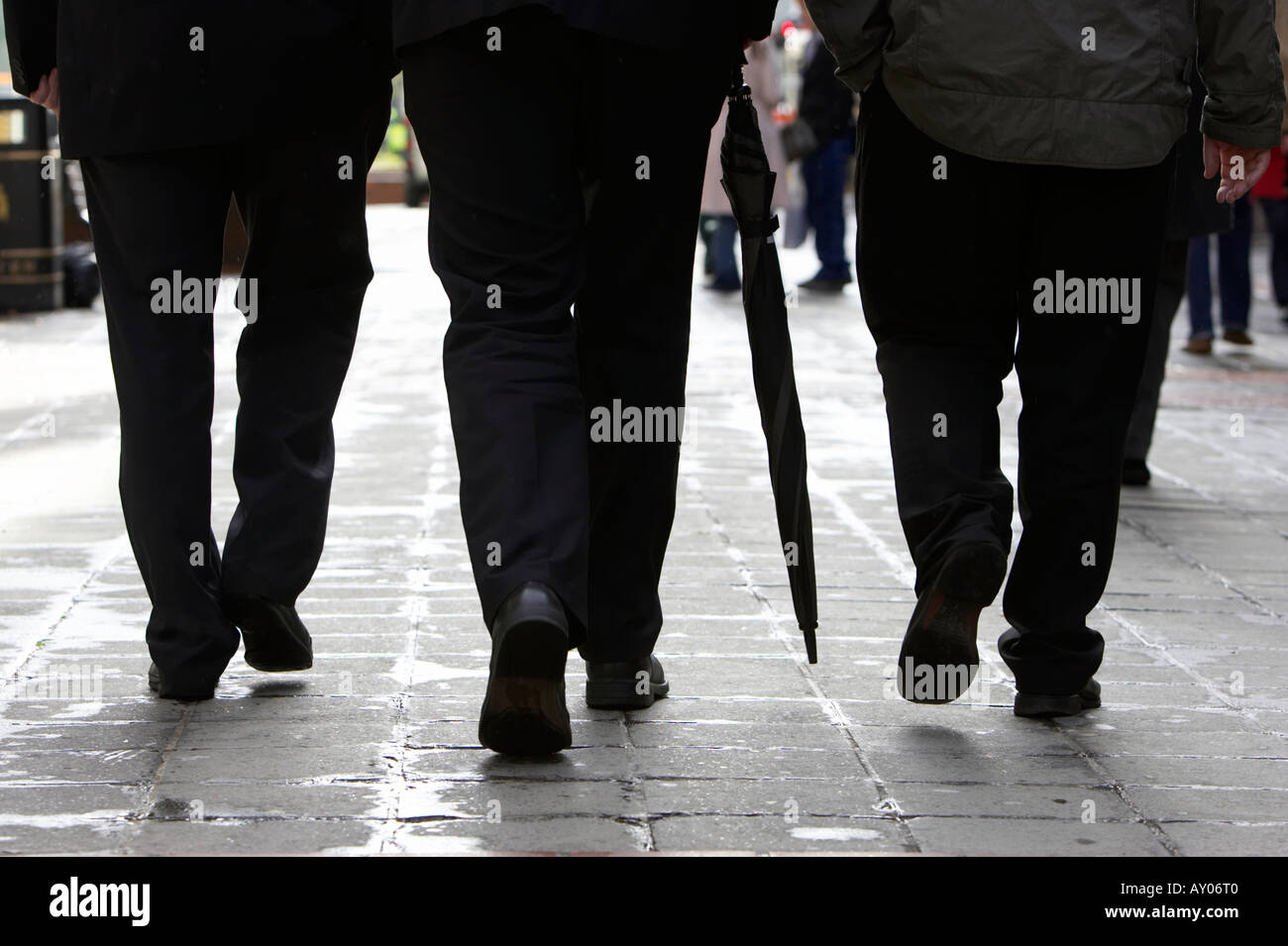 Tre uomini di affari in scuro tute aziendali uno con ombrellone camminando sul marciapiede bagnato in area pedonale a Belfast City Centre Foto Stock