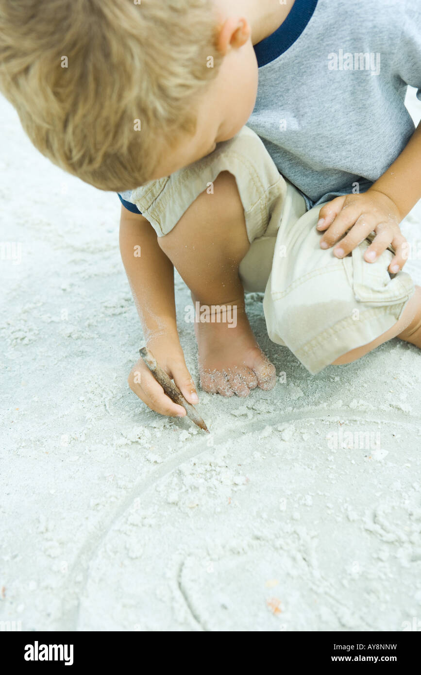 Little Boy accovacciato in spiaggia, disegno in sabbia con stick, testa in appoggio sul ginocchio Foto Stock