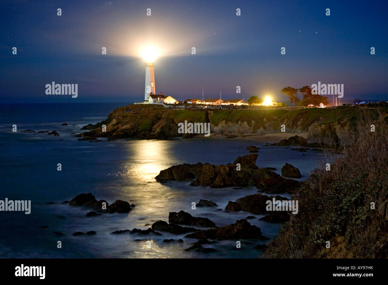 I fasci di luce intorno al centro storico di Pigeon Point Lighthouse al crepuscolo lungo la California s San Mateo litorale con le rocce in primo piano Foto Stock