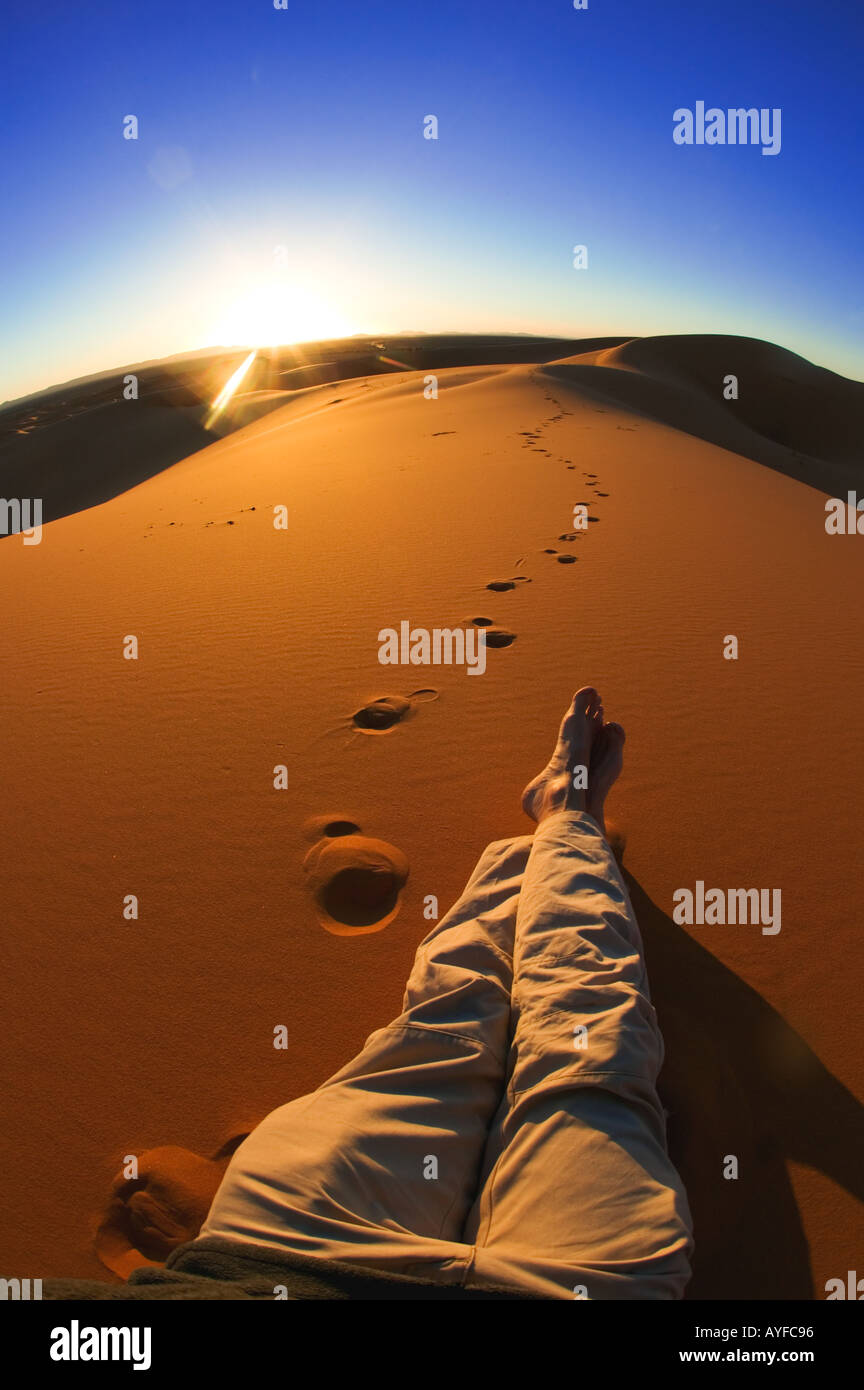 Turismo le gambe della persona rilassarsi e guardare il tramonto nelle dune di sabbia di Erg Chebbi zona deserto del Sahara in Marocco Modello rilasciato Foto Stock