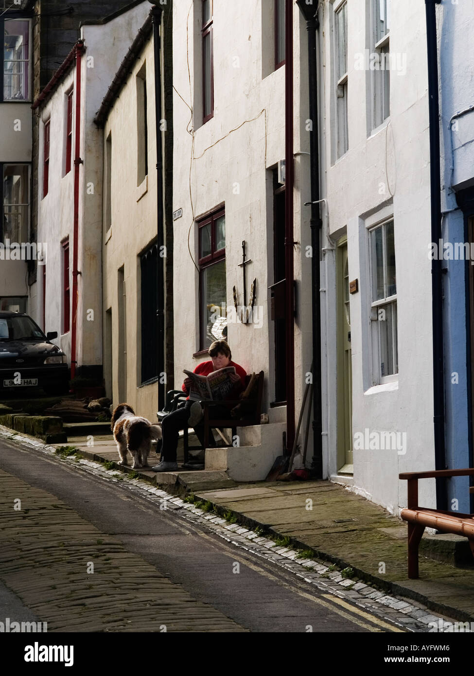 Una donna con un cane si siede al sole leggendo un giornale della domenica al di fuori di un cottage in Staithes North Yorkshire Regno Unito Foto Stock