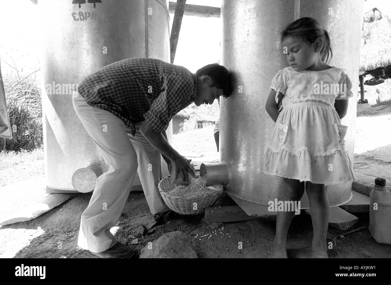 B/W di un agricoltore con sua figlia, che attinge mais dai loro piccoli sili di grano di metallo e di prova di peste in una piccola fattoria di famiglia in Honduras rurale. Foto Stock