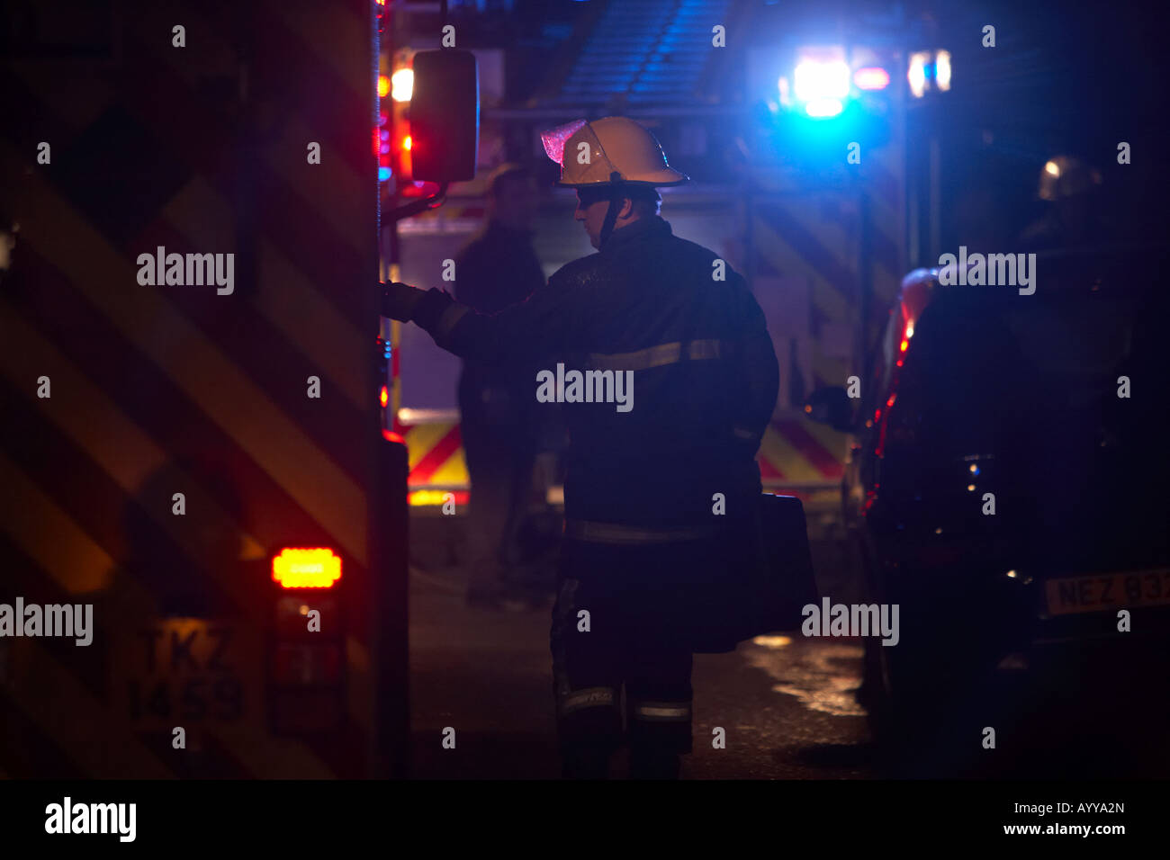 Vigile del fuoco che indossa un casco e tuta protettiva che trasportano un caso apre le porte di un motore fire su una strada di notte Irlanda del Nord servizio antincendio Foto Stock