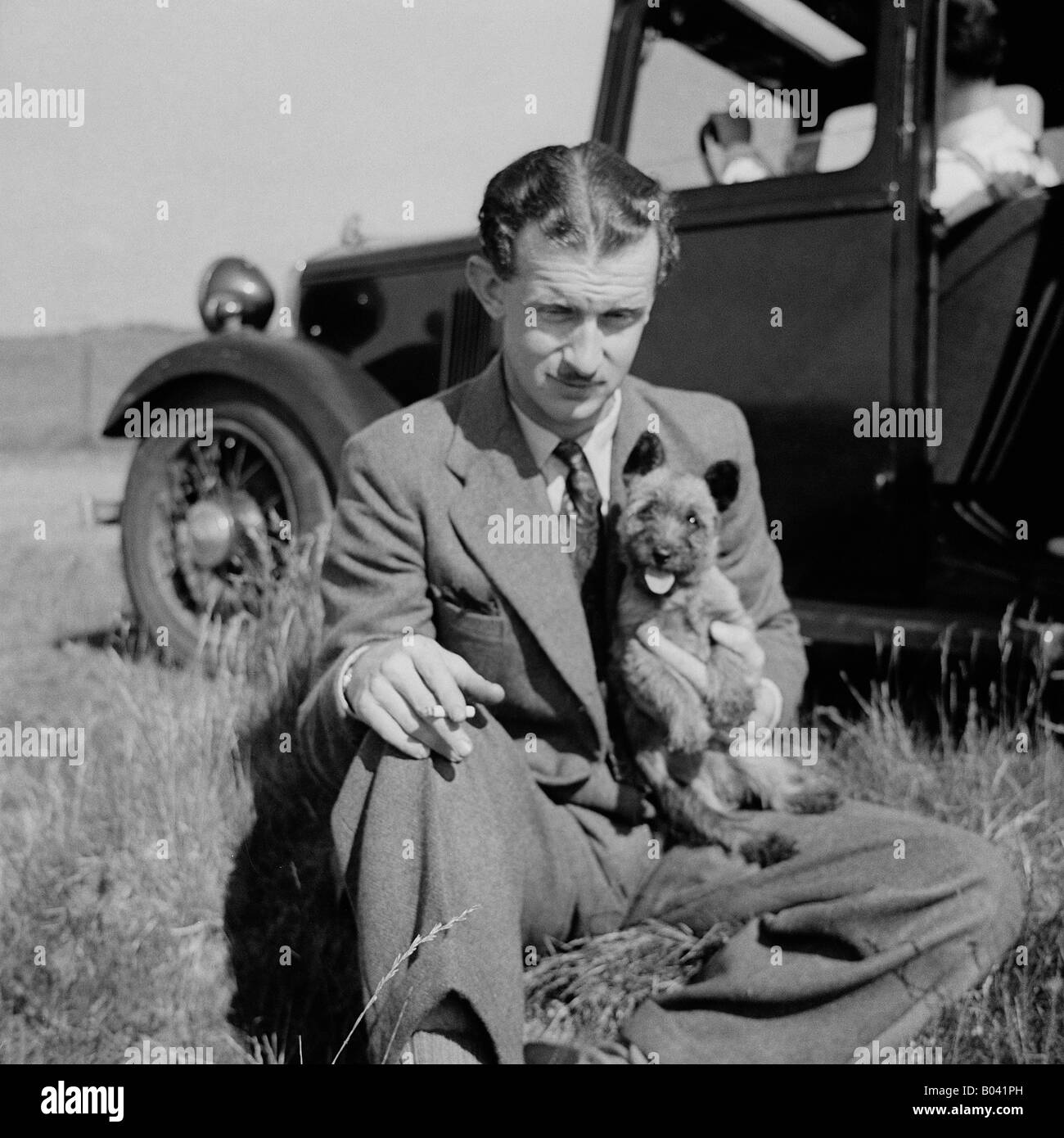 Vecchio vintage snapshot dell uomo seduto nel campo Azienda piccolo cane CON AUTO D'EPOCA IN BACKGROUND Foto Stock